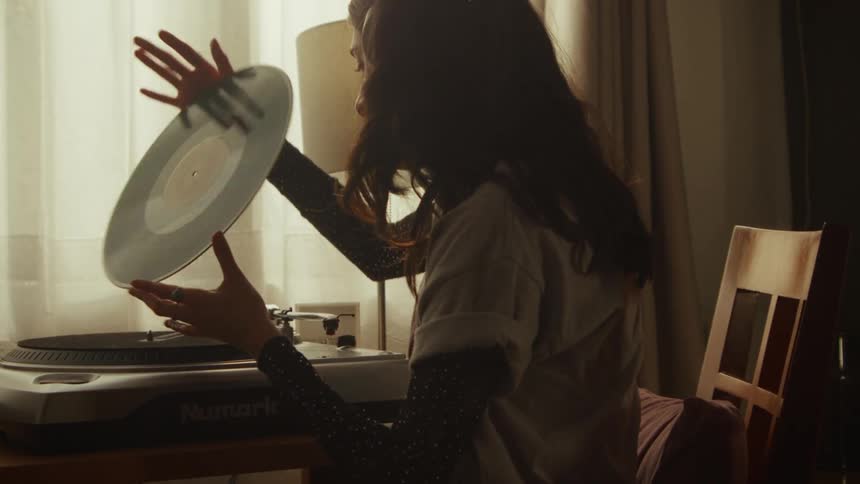In a cozy room with soft lighting, a woman carefully places a vinyl record on a turntable in slow motion