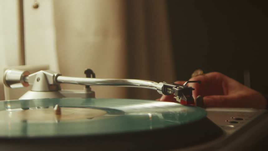 A hand carefully places the needle on a spinning teal vinyl record in slow motion on a vintage turntable
