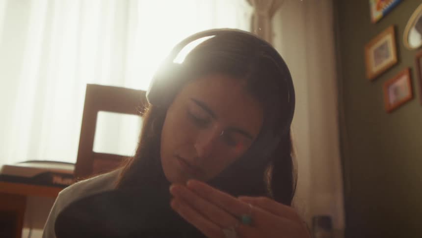 In a warmly lit room, a woman with headphones on listens to music and affectionately holds a vinyl record