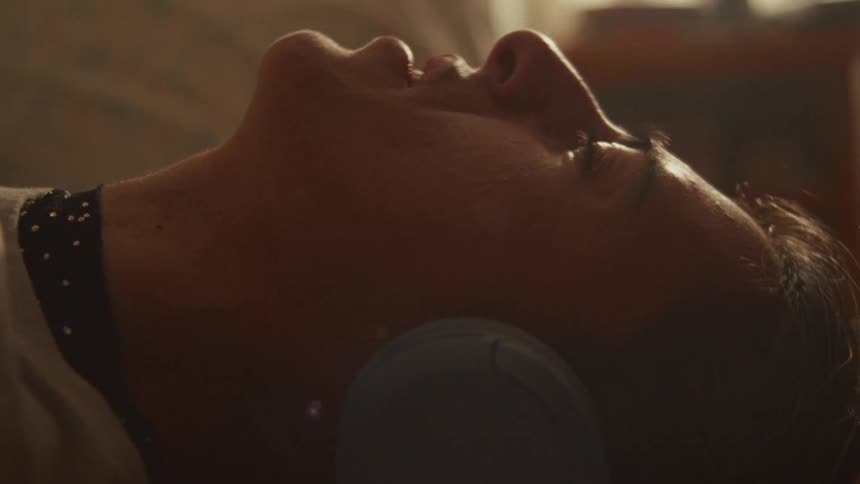 A close-up of a man with his eyes closed, lying down and peacefully listening to something on his headphones in warm, gentle lighting