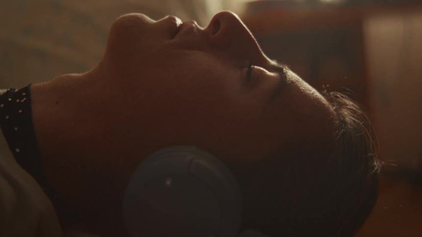 A young woman peacefully lies down, listening to music on her headphones as dust motes dance in the warm light