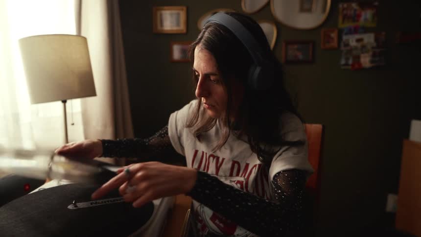 A young woman wearing headphones carefully places a vinyl record on a turntable in her cozy room