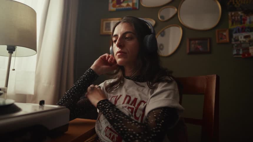 A pensive young woman in her moody bedroom puts on headphones to listen to a record, her expression becoming contemplative