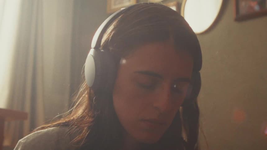 A young woman with her eyes closed peacefully listens to music on headphones in a hazy, sunlit room