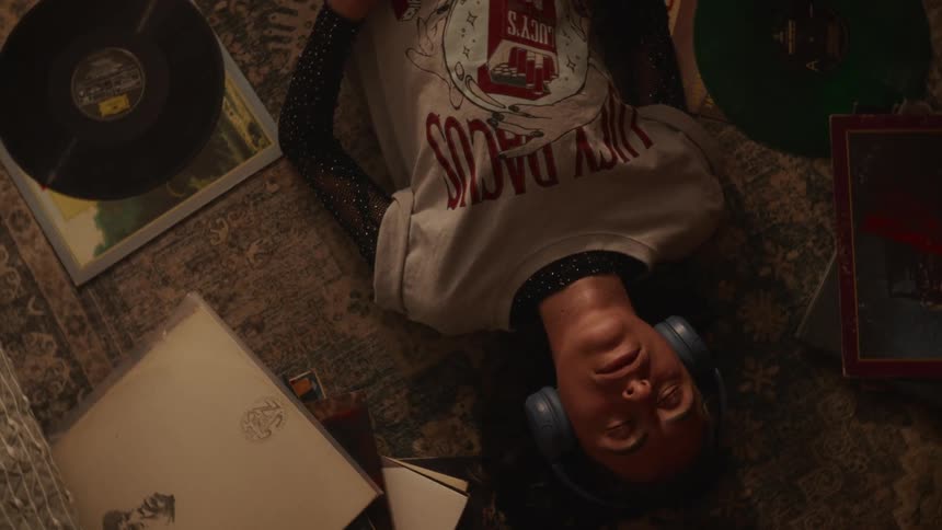 An overhead shot of a young woman with headphones on, peacefully lying on the floor surrounded by her vinyl record collection