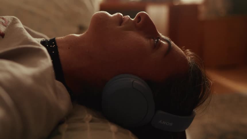 A close-up profile of a young woman peacefully lying down, listening to audio on wireless headphones in a warm, cozy room