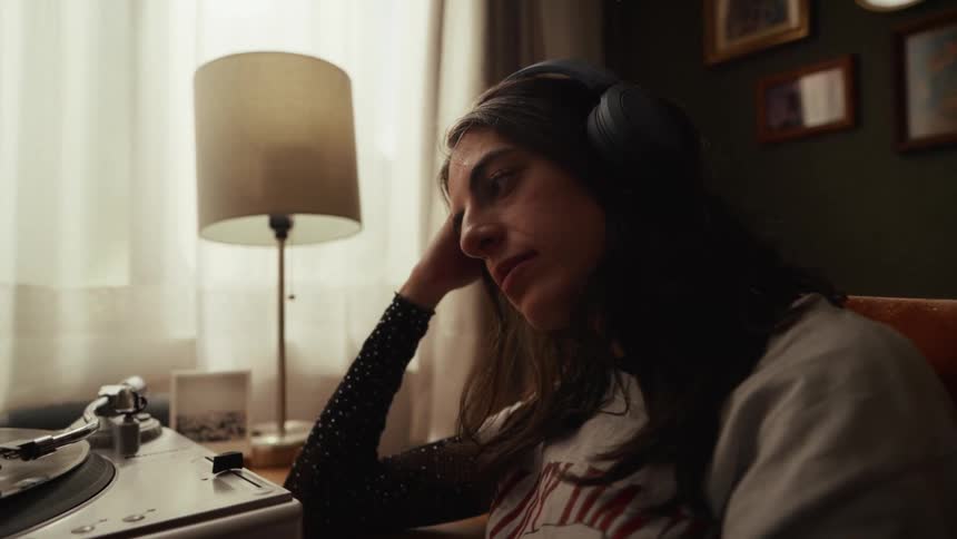 A close up of a sad young woman wearing headphones while listening to music from a record player in her room