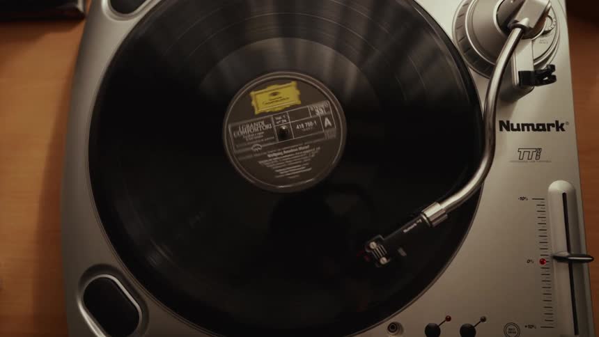 A top-down view shows a hand lowering the needle onto a classic vinyl record as it begins to play on a turntable