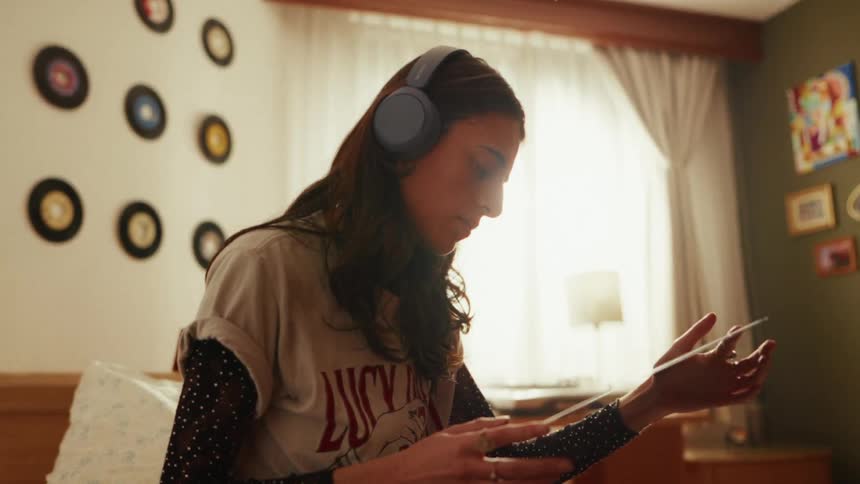 A young woman with headphones on sits in her bedroom and looks at a vinyl record she's holding