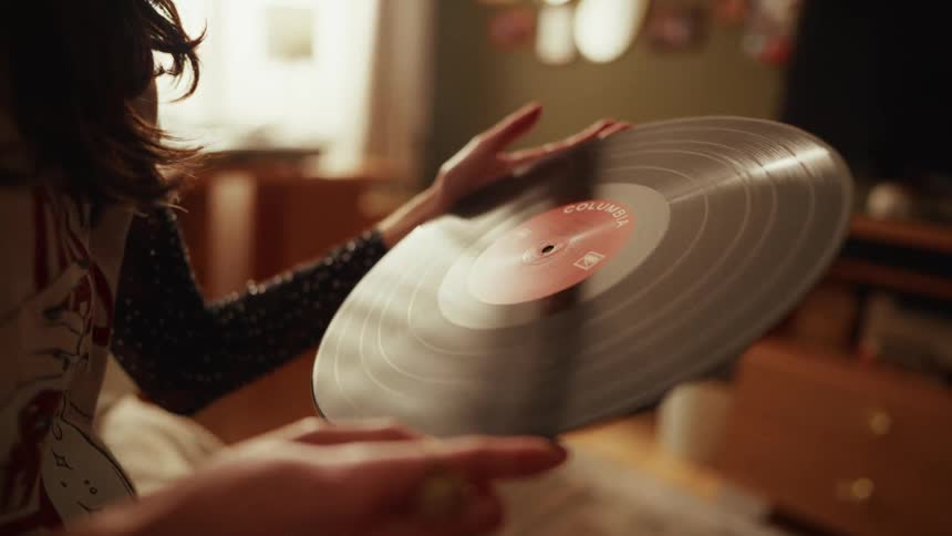 A woman holds and carefully inspects a classic vinyl record in a warmly lit, cozy room