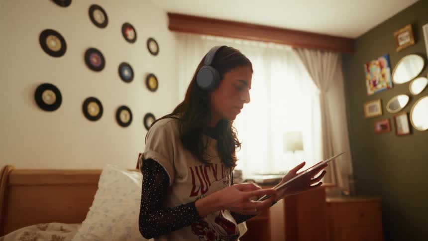 A young woman wearing headphones enjoys her music hobby, examining a vinyl record while using a tablet in her cozy, retro-decorated bedroom