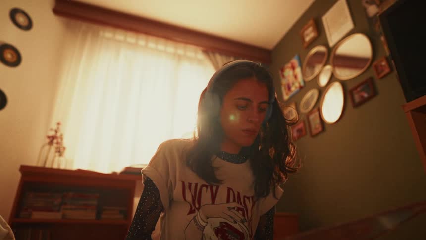 A nostalgic young woman wearing headphones enjoys music while looking at a vinyl record cover in her sunlit retro-style bedroom