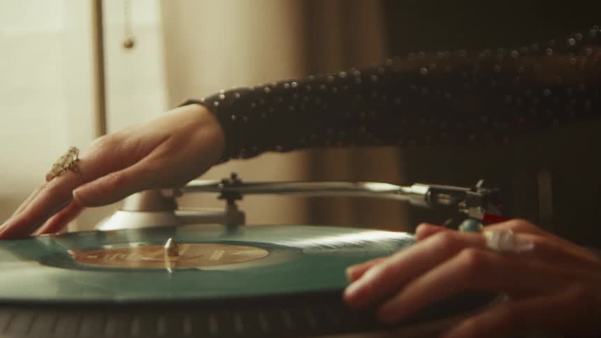 A person wearing rings and a sparkly shirt carefully places a teal vinyl record on a turntable and sets the needle to play