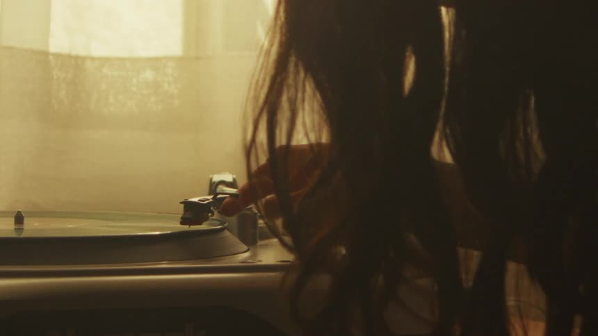A woman carefully places the needle on a spinning record on a vintage turntable in a warmly lit room