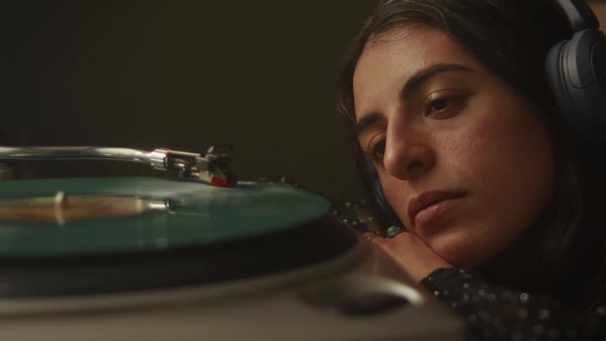 A pensive woman in a dimly lit room listens intently to a vinyl record playing on a turntable