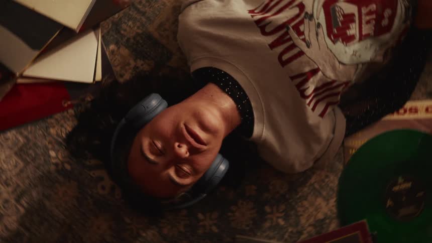 Top-down rotating shot of a relaxed person with headphones on, eyes closed, enjoying music while lying on a carpet surrounded by vinyl records
