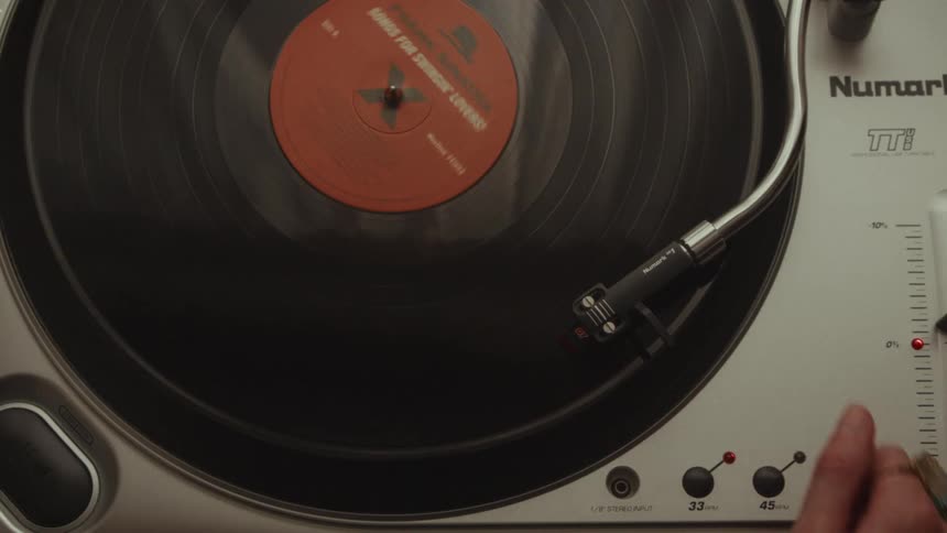 A high-angle shot captures a hand placing a classic vinyl record onto a turntable, starting it up, and dropping the needle to play the music