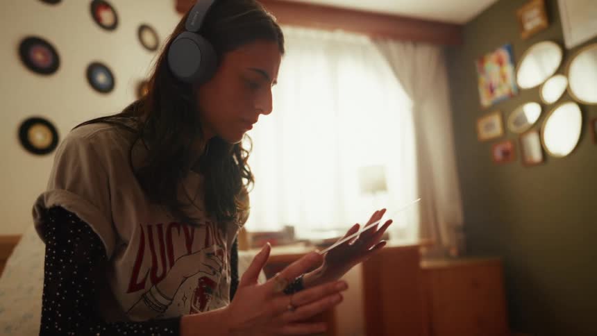 A young woman wearing headphones enjoys a quiet moment in her warmly lit bedroom, carefully examining a vinyl record from her collection