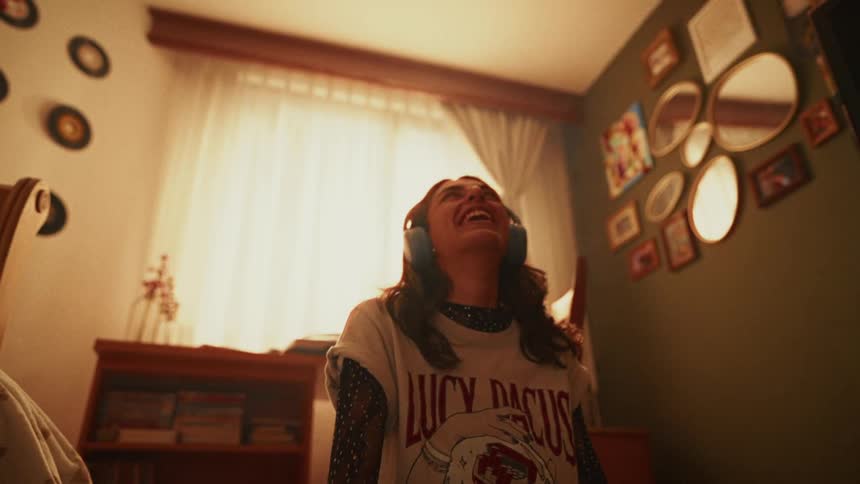 A carefree young woman wearing headphones bursts into laughter in slow motion in her warmly lit, decorated bedroom
