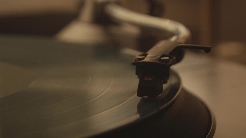 A close-up shot shows a record player's needle tracing the grooves of a spinning vinyl record in warm, ambient light