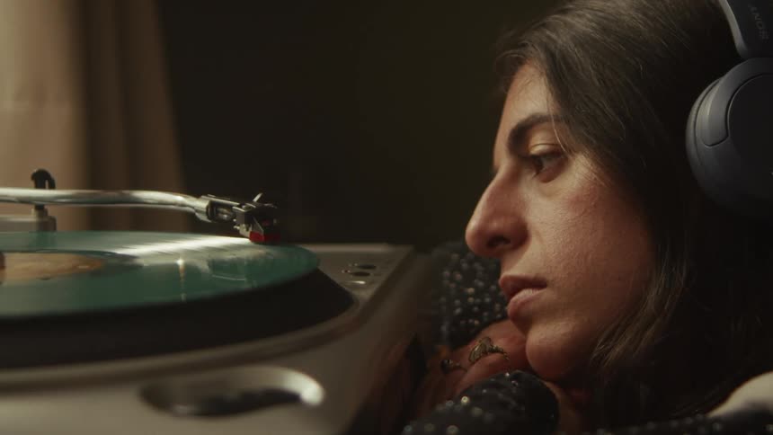 A woman in headphones listens to a spinning vinyl record on a turntable in a dimly lit, cozy room