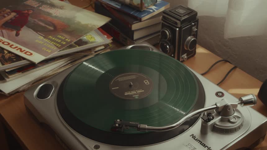A person with rings on their fingers carefully places a green vinyl record on a turntable to play music in a retro room