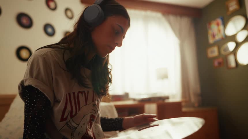 A young woman wearing headphones carefully inspects a vinyl record while sitting in her cozy, retro-decorated bedroom