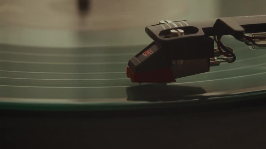 A close-up, macro shot shows a finger carefully lifting the needle of a tonearm from a spinning green vinyl record and placing it back down again