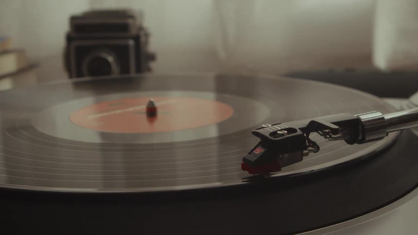 A close-up of a vinyl record playing on a turntable, with a vintage camera out of focus in the background