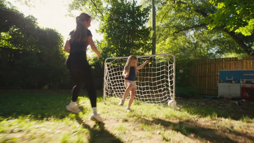 A mother scores a slow-motion goal on her daughter while playing soccer together in their sunny backyard