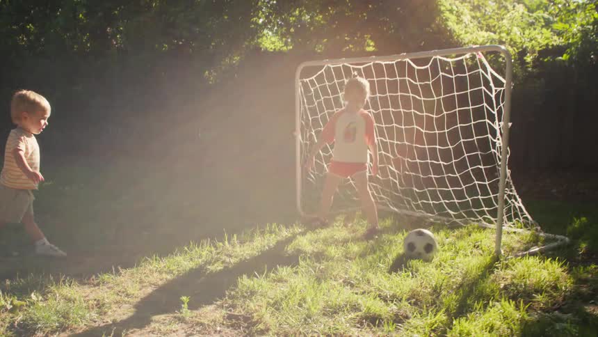 A toddler boy kicks a soccer ball to his sister who is playing goalie in their sunny backyard