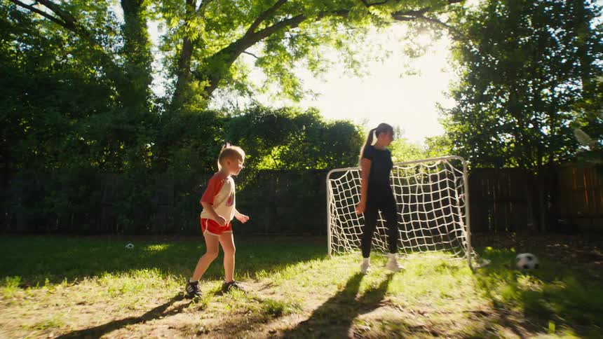 A young girl scores a soccer goal against her sister in a sun-drenched backyard during a playful afternoon game