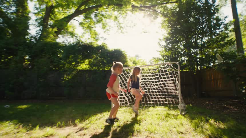 Two young girls play a game of soccer in a sunny backyard, with one taking a shot on goal as the other defends