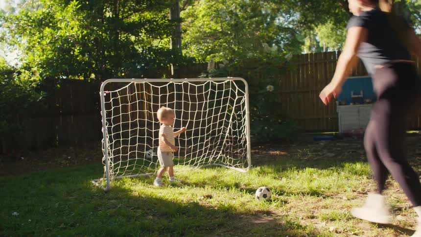 A happy family enjoys a playful game of soccer in their sunny backyard, with a toddler acting as the goalie