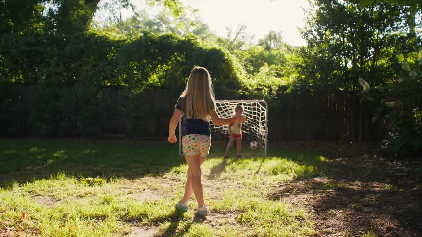 A young girl scores a soccer goal against her brother in their sunny backyard and smiles happily at the camera