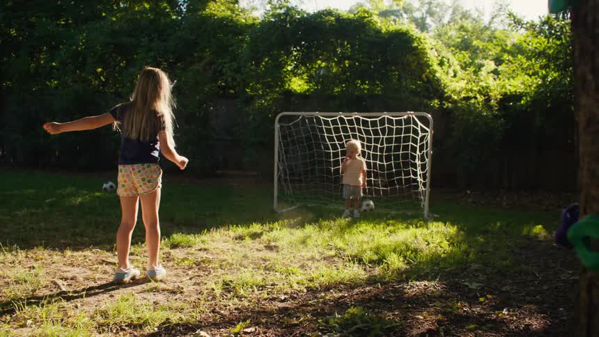 A big sister playfully kicks a soccer ball to her little brother standing in a goal in their sunlit backyard