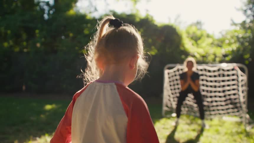 A young girl in a ponytail kicks a soccer ball towards the goal where her sister is playing keeper in their sunny backyard