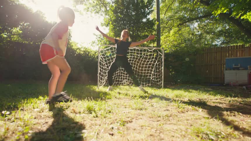 A young girl scores a goal while playing soccer with her mom in their sunny backyard and celebrates in joy