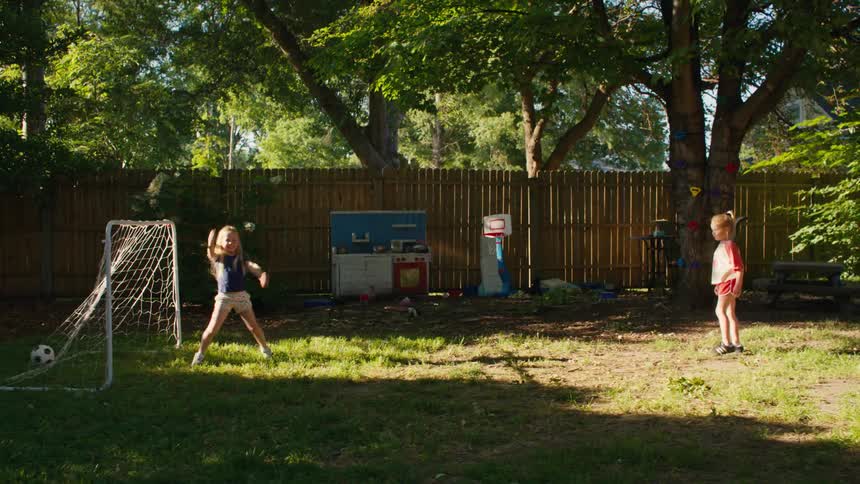 A little girl scores a soccer goal on her sister in the backyard, who then playfully celebrates while catching bubbles