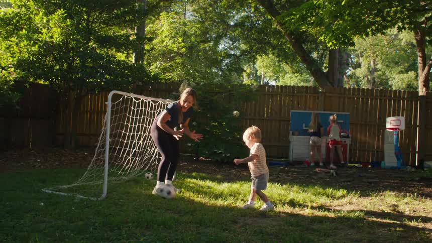 A toddler scores a goal on his mom in their backyard, and she celebrates with a clap and a high five
