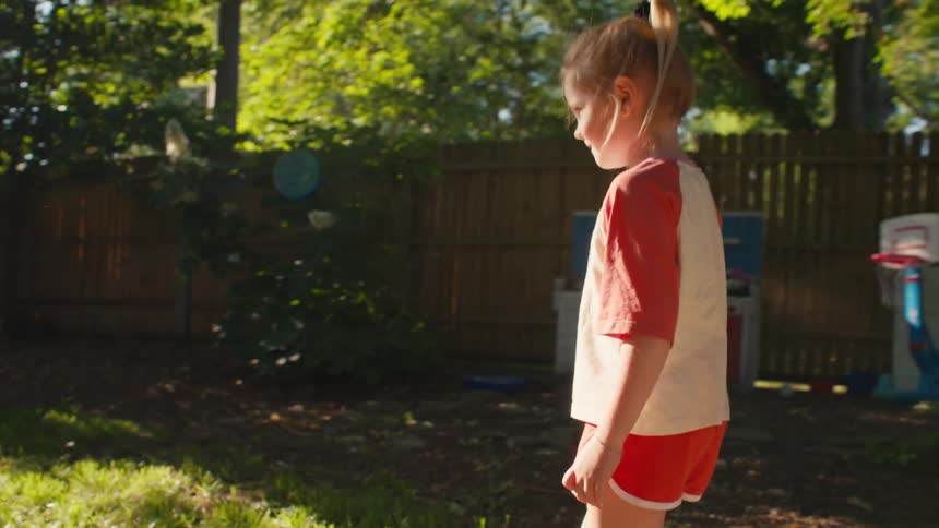A little girl in red shorts kicks a soccer ball towards her sister playing goalie in their sunlit backyard