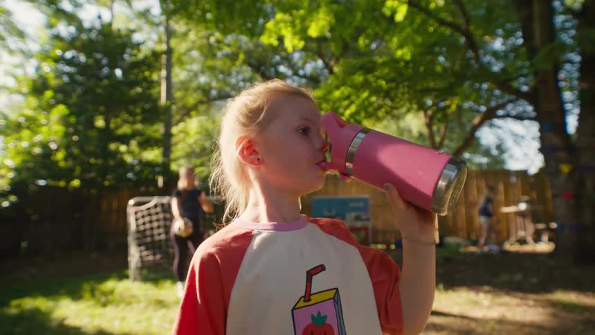 A young girl takes a refreshing drink from her pink water bottle while playing in the backyard on a sunny day