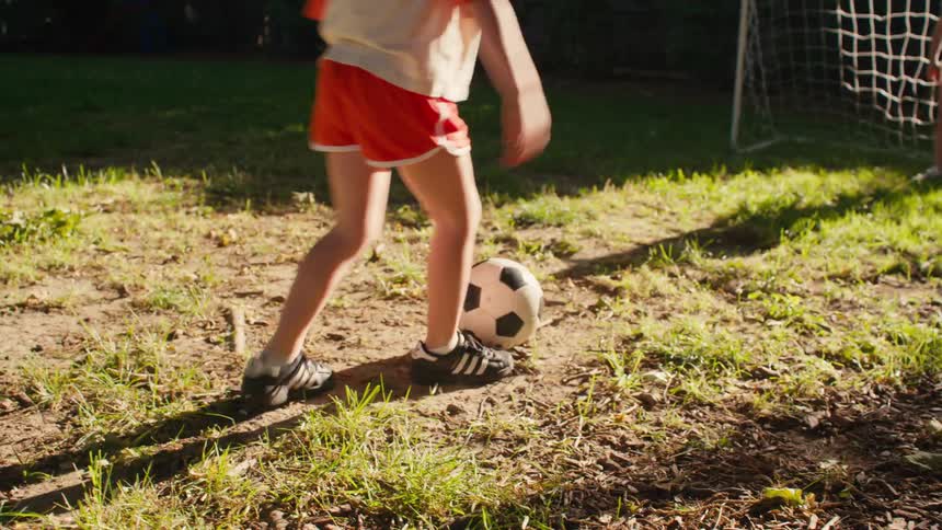 A young girl kicks a soccer ball as her friend guards the goal, and the goalie celebrates the goal with a happy dance in their sunny backyard