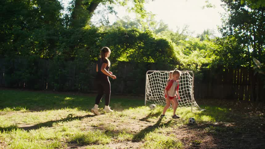 A mom and her two young daughters have fun playing soccer together in their sunlit backyard