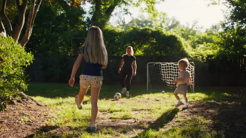A young girl dribbles a soccer ball while playing with her mom and toddler brother in their sunny backyard
