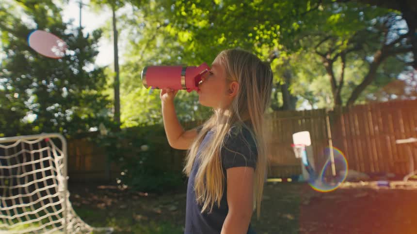 A young blonde girl takes a refreshing drink from her pink water bottle in a sun-drenched backyard playground