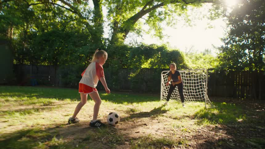 A young girl kicks a soccer ball and scores a goal against her mother playing goalie in their sunny backyard