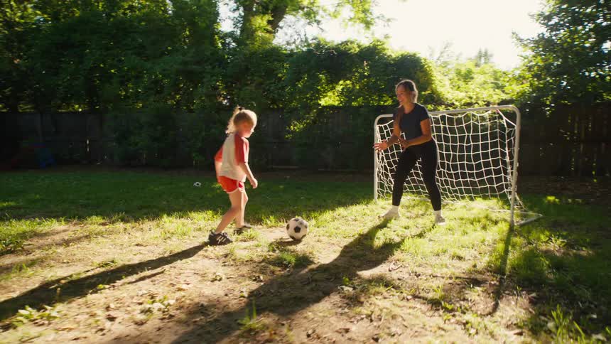 A little girl scores a goal while playing soccer with her mom in the backyard and happily jumps for joy to celebrate