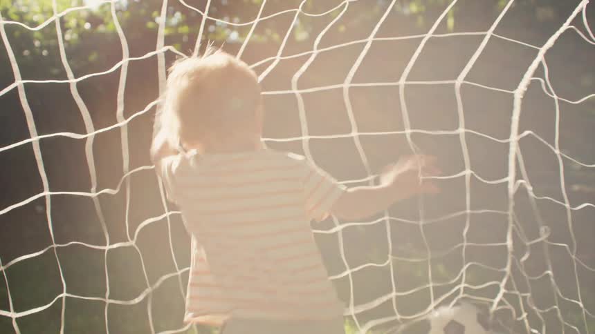 A cute toddler stands as goalie in a sunlit backyard, then joyfully runs out of the goal before playfully tumbling onto the grass