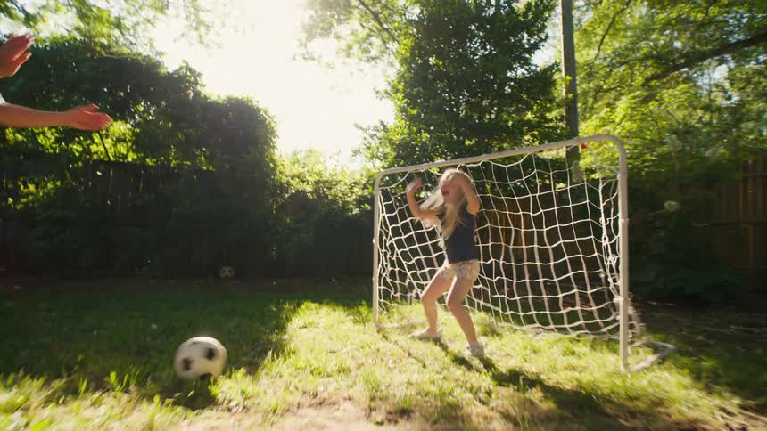 A mother playfully practices soccer with her young daughter who successfully stops the ball, and they celebrate with a high-five in the sunny backyard